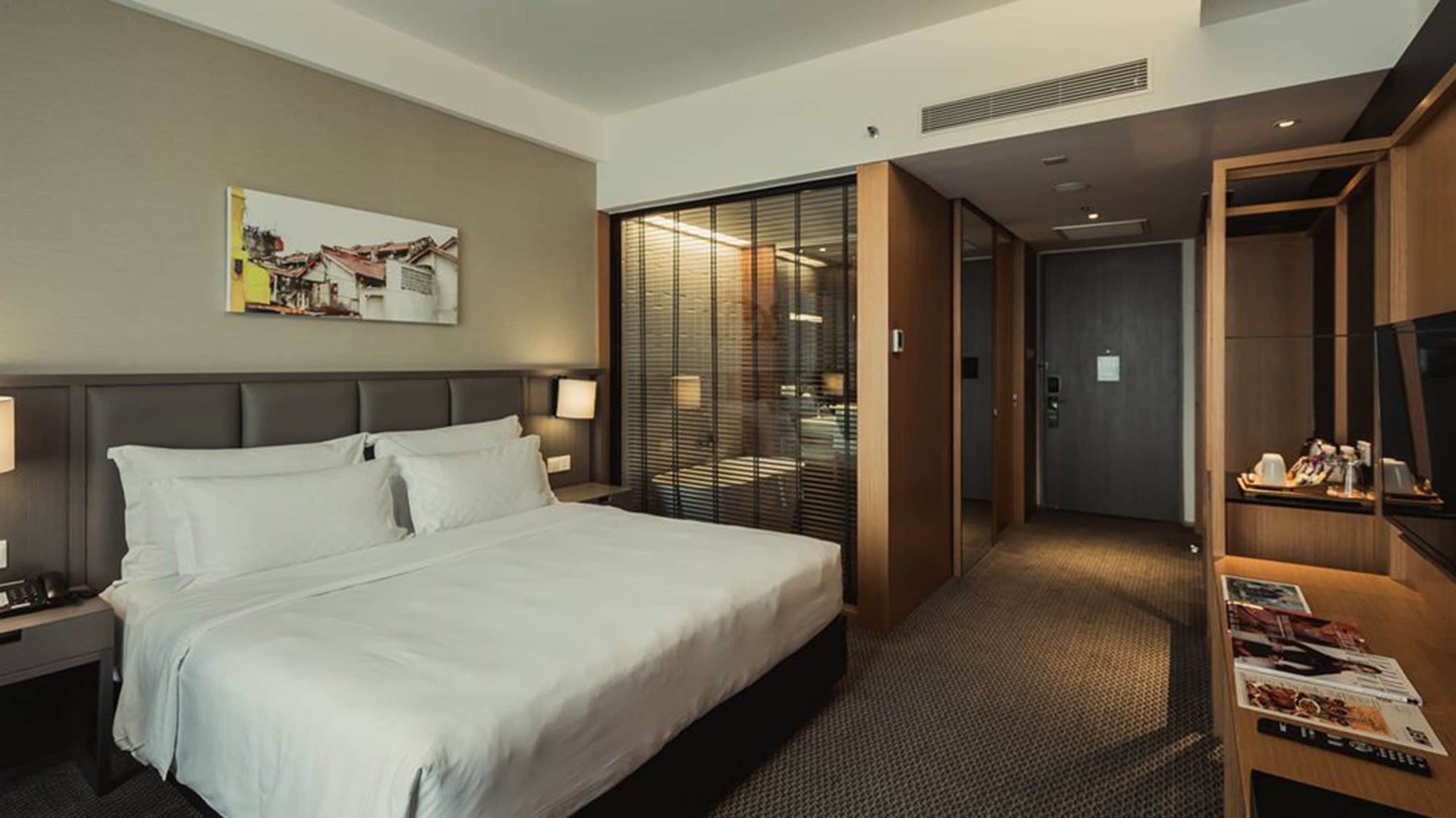 A hotel room with a large bed, a nightstand, a bedside table, a wall-mounted lamp, a painting, a sliding door leading to a bathroom, and a small desk with a tray of items.