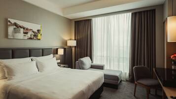 A hotel room with a large bed, a desk, a chair, a lamp, and a window with curtains.