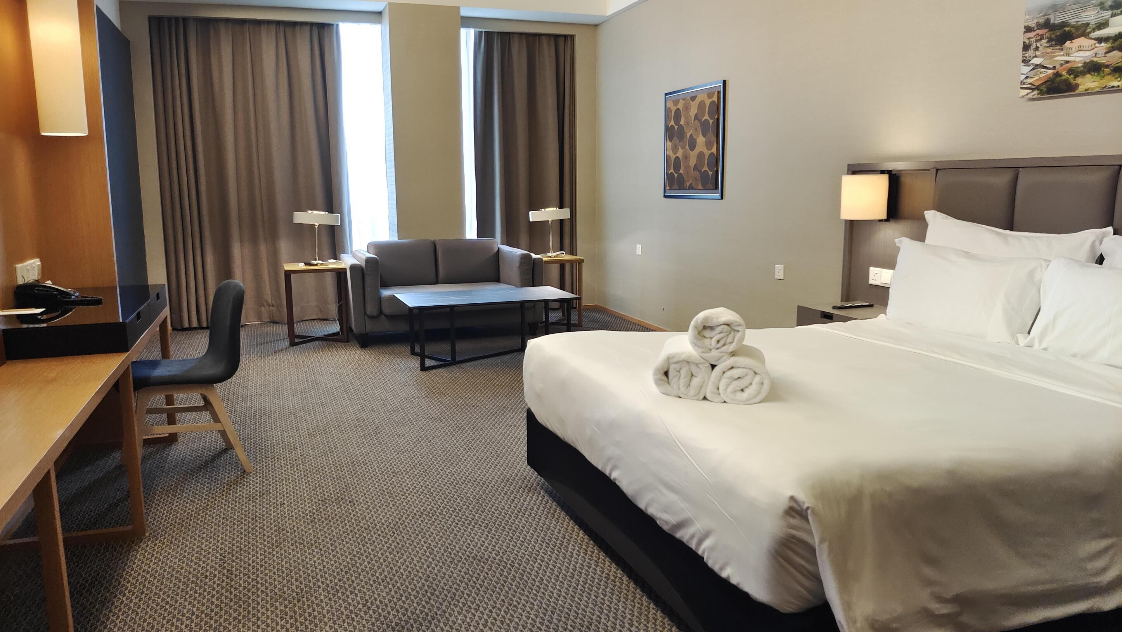 A hotel room with a bed, a desk, a chair, a small table, and a lamp.
