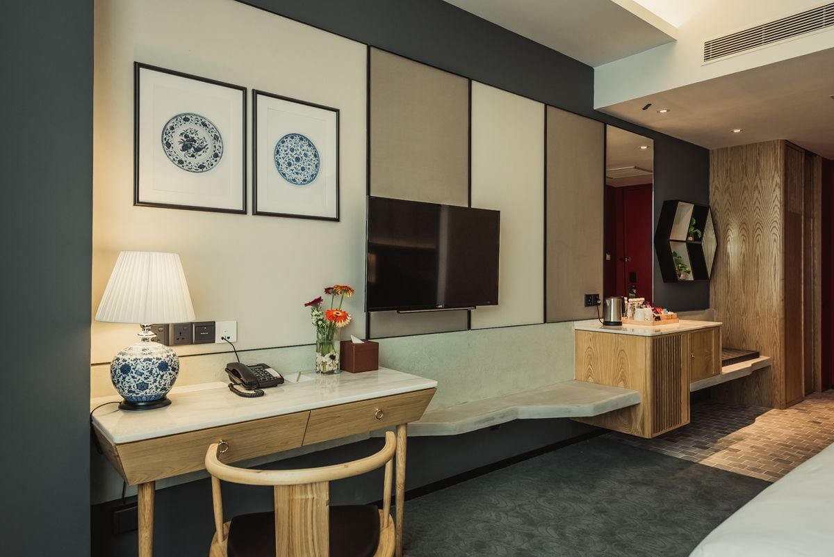 A modern hotel room with a desk, chair, bedside table, TV, and a mirror.