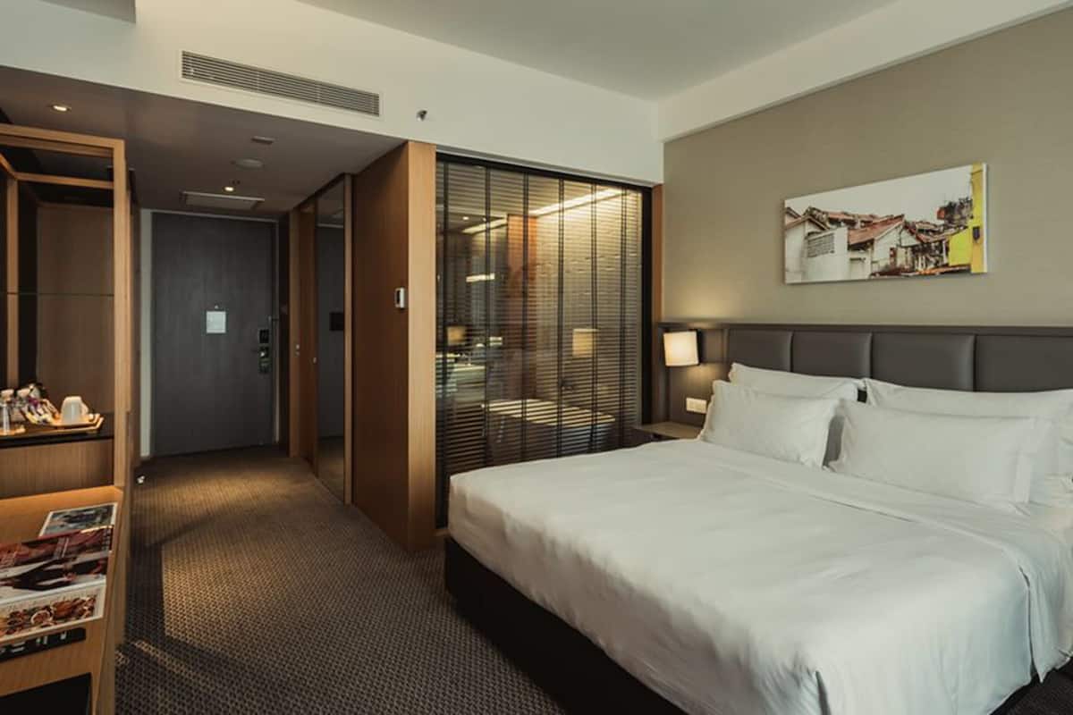 A hotel room with a large bed, a desk with a computer and magazines, a minibar, and a sliding door leading to a bathroom.