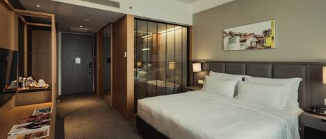 A hotel room with a large bed, a desk with a computer and magazines, a minibar, and a sliding door leading to a bathroom.