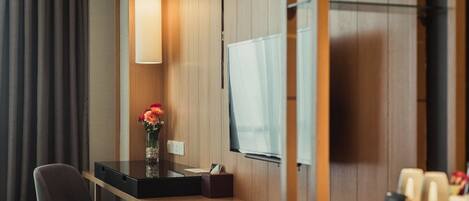 A modern hotel room with a wooden panel wall, a desk with a chair, a vase with flowers, a flat-screen TV, and a coffee station.