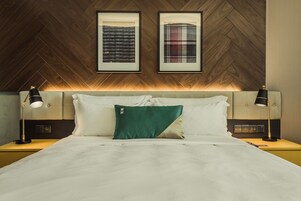 Una habitación de hotel con techo de madera en patrón de espiga, una cama con ropa de cama blanca, dos mesitas de noche con lámparas y dos cuadros en la pared.