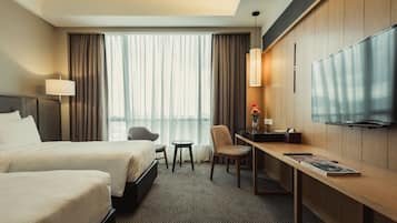 Ein Hotelzimmer mit zwei Betten, einem Schreibtisch, einem Sessel, einem Fernseher und Blick auf die Stadt durch große Fenster.