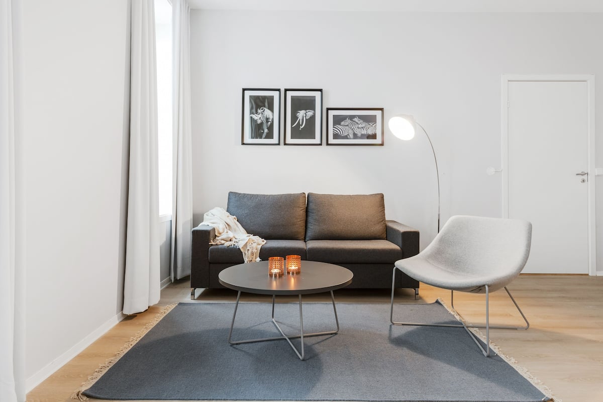 A modern living room with a grey sofa, a round coffee table, and a white chair. There are three framed pictures on the wall and a floor lamp.