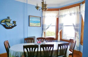 A dining room with a round table, wooden chairs, a chandelier, a painting, and curtains. - The Harbor House Bed & Breakfast (Staten Island)