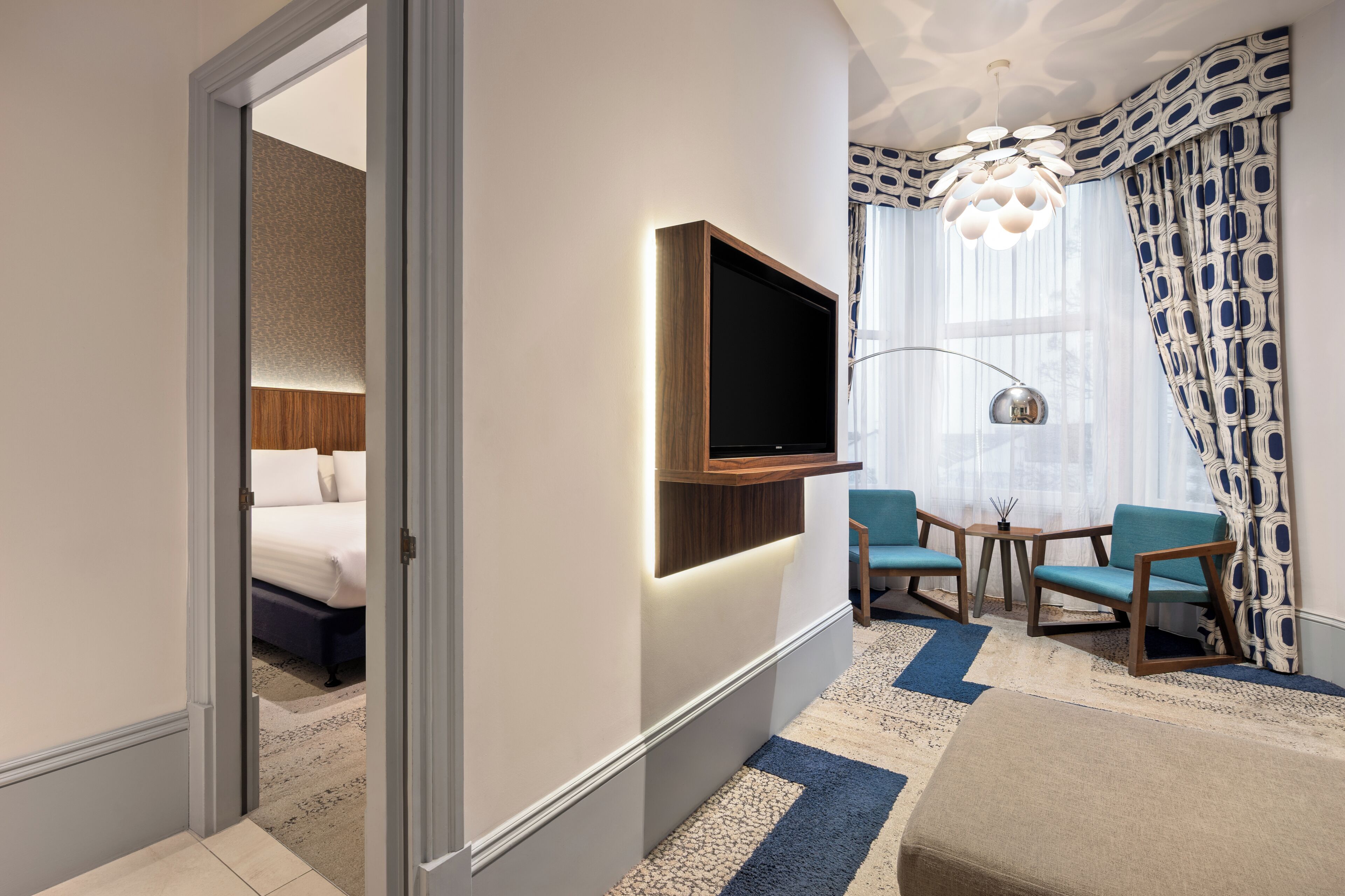 A hotel room with a bed, a TV mounted on the wall, two chairs, a small table, and a patterned rug.