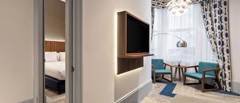 A hotel room with a bed, a TV mounted on the wall, two chairs, a small table, and a patterned rug.