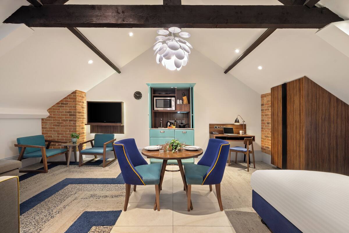 Een moderne loft slaapkamer met zichtbare balken, een zithoek met blauwe stoelen, een keuken met een turquoise kast, en een groot bed.