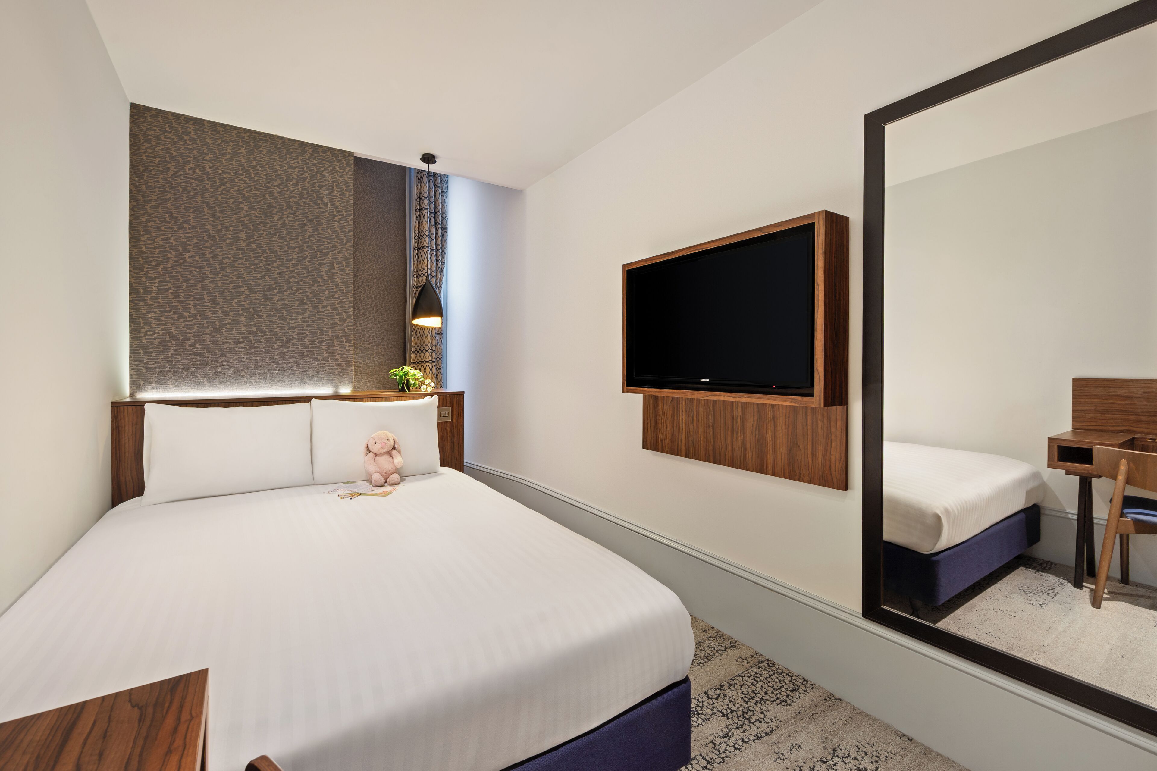 Una habitación de hotel moderna con una cama grande, un televisor de pantalla plana montado en la pared, una mesita de noche con lámpara y un espejo amplio.