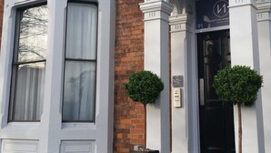 Un edificio en esquina con ventanal saliente, puerta negra y un pequeño árbol al frente.
