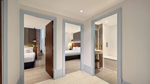 A hotel room with a bed, bedside table, lamp, and a view of another room.