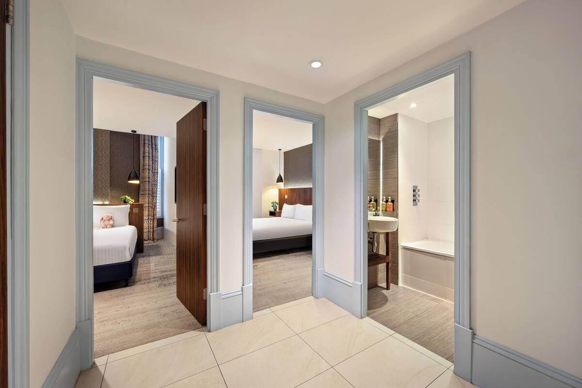 Een hotelkamer met een bed, nachtkastje, lamp en uitzicht op een andere kamer.