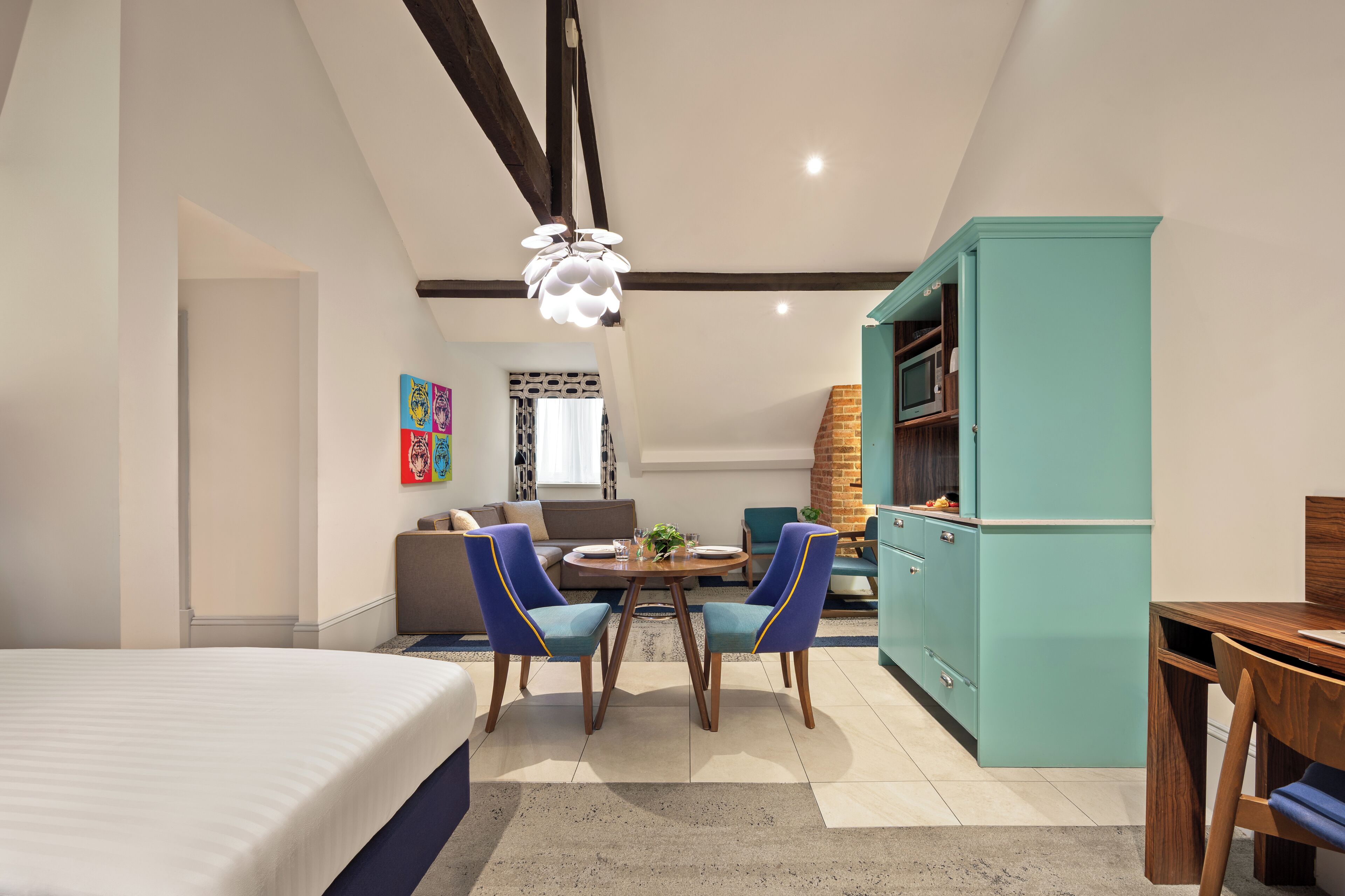 A modern hotel room with a bed, a dining area with chairs and a table, a kitchen with cabinets, and a living area with a sofa and a TV.