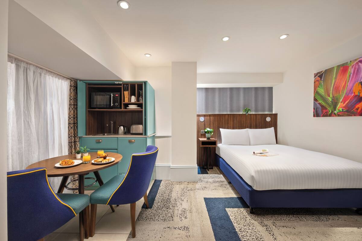 Een compacte hotelkamer met een bed, een eettafel met stoelen, een kitchenette en een klein nachtkastje met een plant.