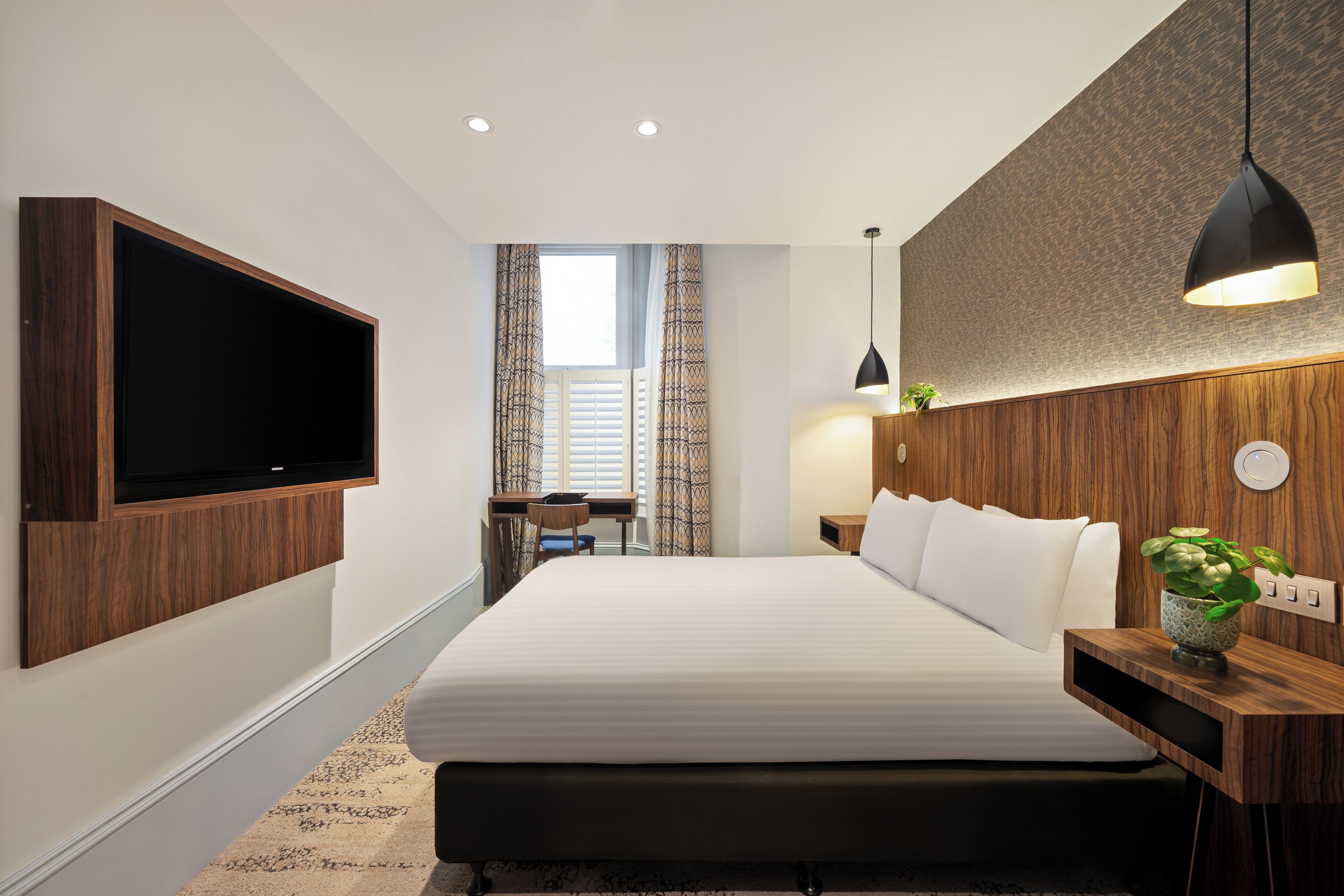 A modern hotel room with a large bed, a flat-screen TV mounted on the wall, a desk with a chair, and a bedside table with a plant.