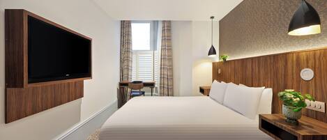 A modern hotel room with a large bed, a flat-screen TV mounted on the wall, a desk with a chair, and a bedside table with a plant.