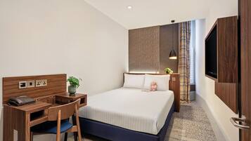 A hotel room with a bed, desk, chair, TV, and a plant.