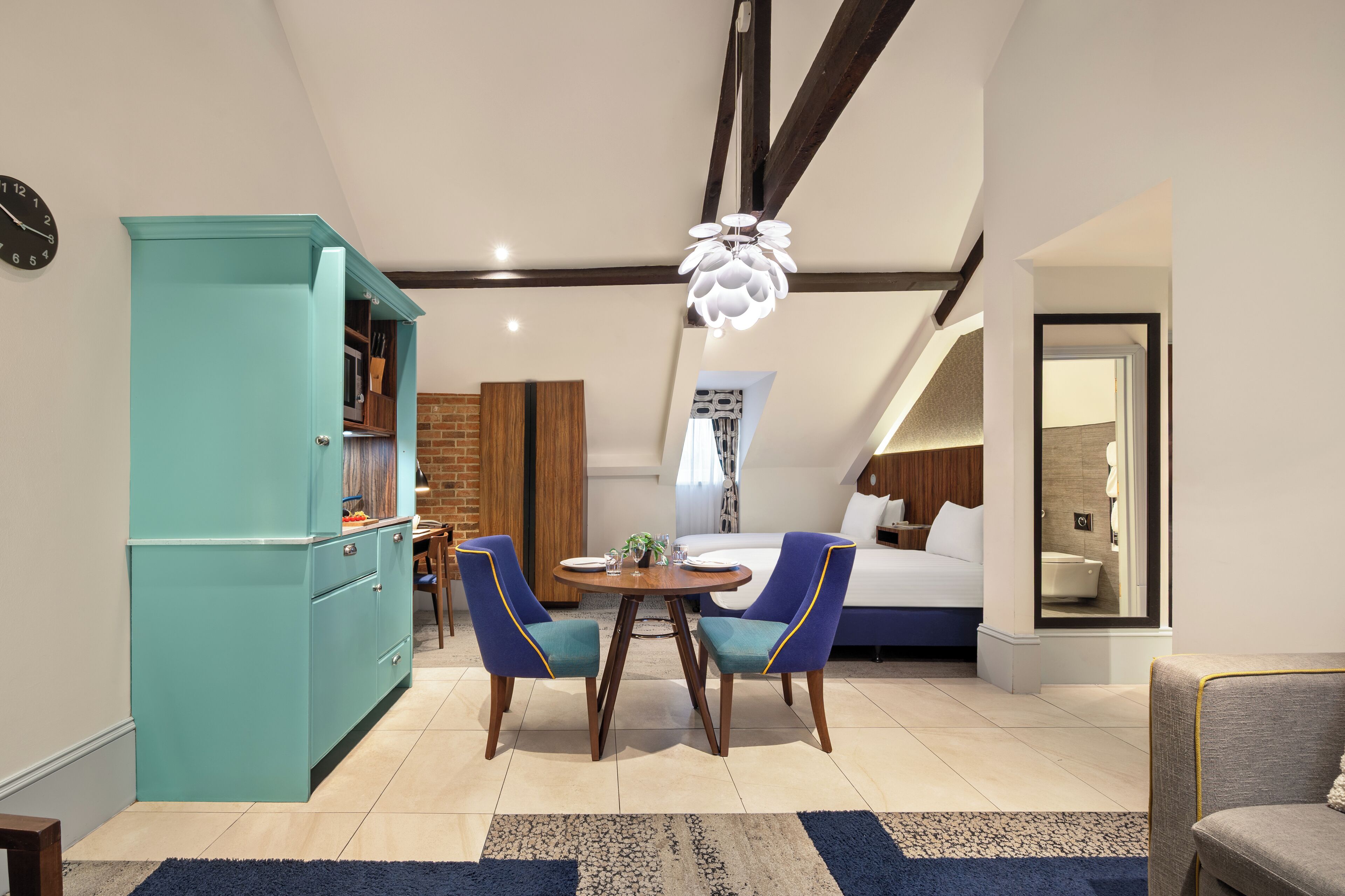 Una habitación moderna con una cama, una mesa de comedor con sillas, una kitchenette y un baño visible a través de una puerta abierta.