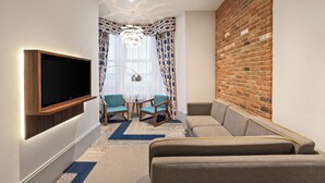 A modern living room with a flat-screen TV, two blue chairs, a sofa, a patterned rug, and a brick accent wall.