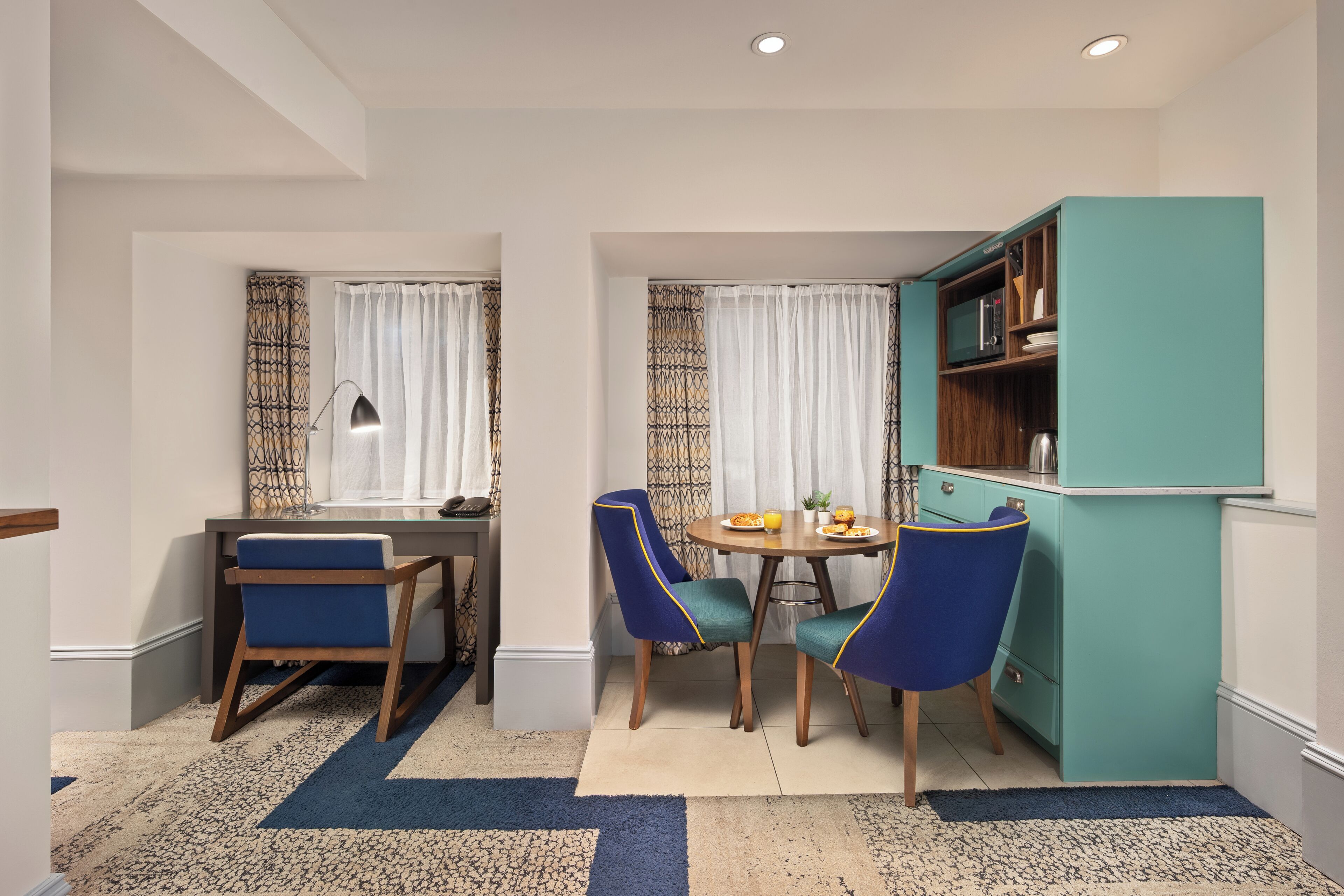 A compact kitchen with a teal cabinet, a dining table with two chairs, and a small refrigerator.