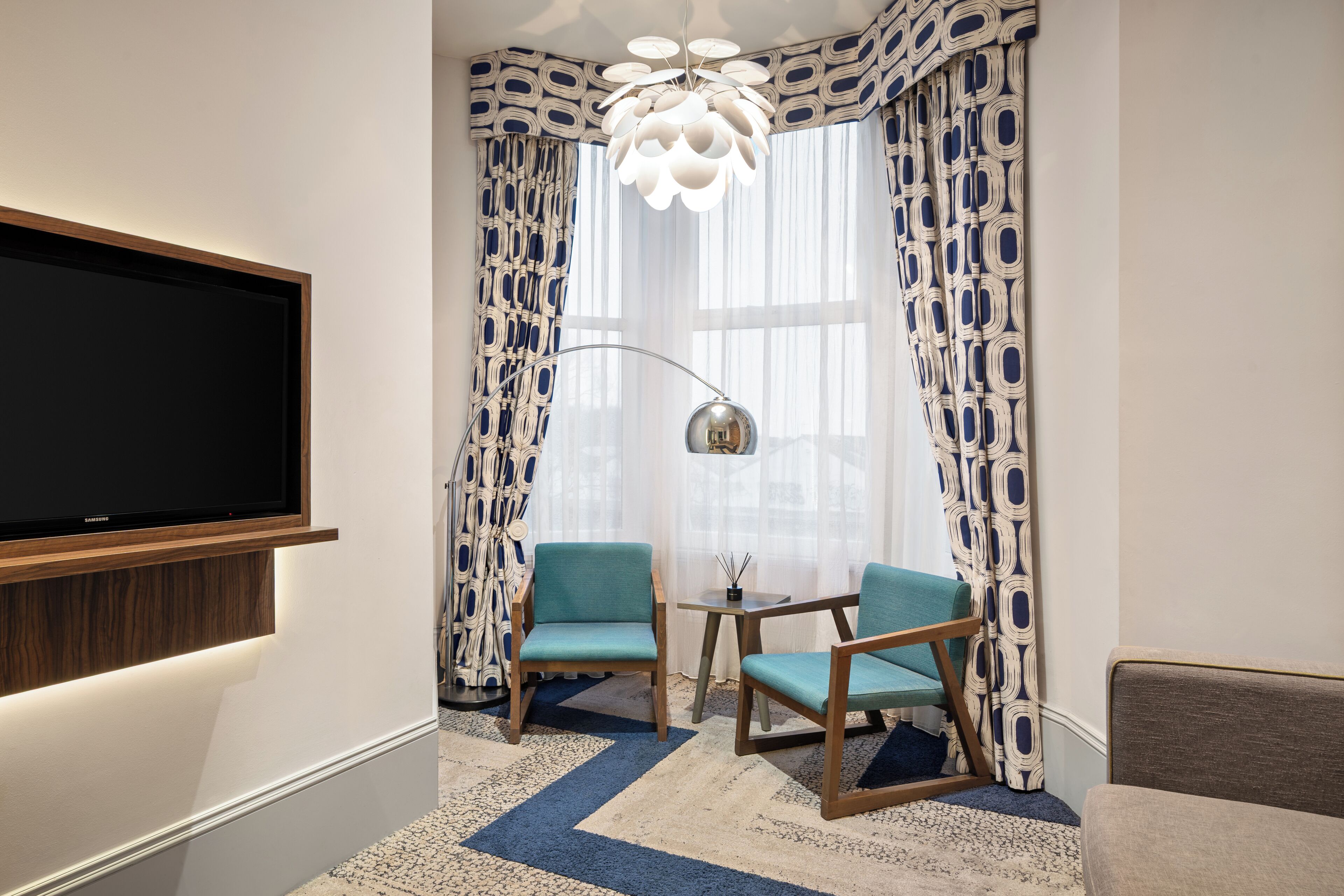 A modern room with a flat-screen TV, two chairs, a small table, and patterned curtains.