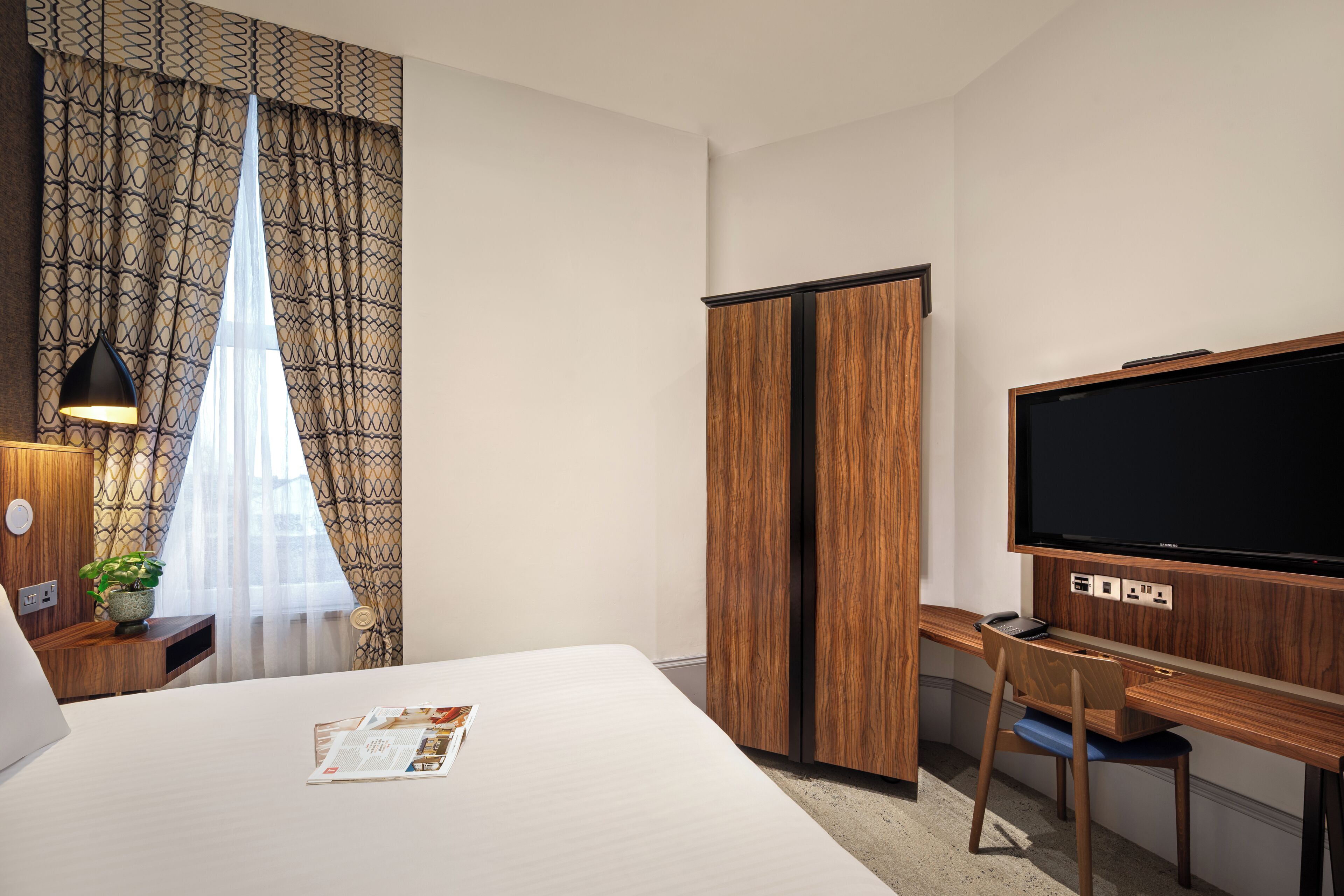 Habitación de hotel con cama, escritorio, televisor, armario y ventana con cortinas.