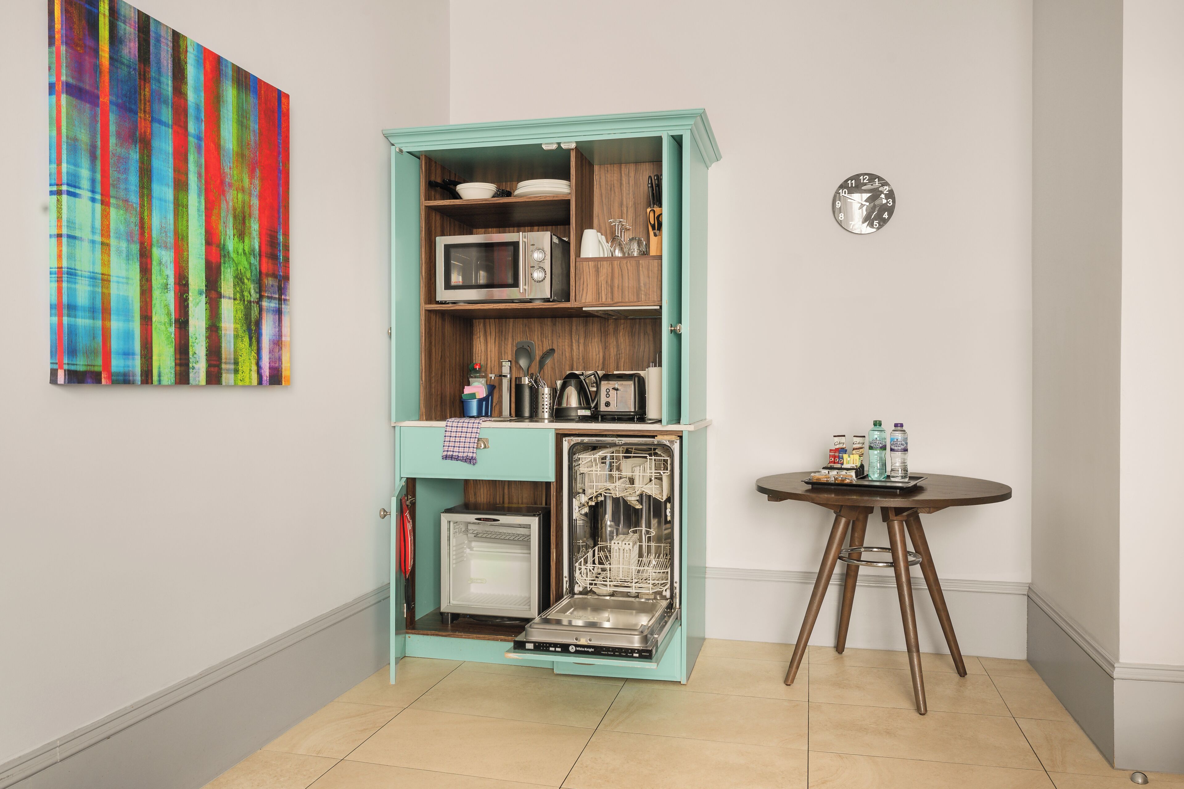 Una cucina compatta con forno a microonde, tostapane e lavastoviglie, un piccolo tavolo con bottiglie e un quadro astratto dai colori vivaci appeso al muro.