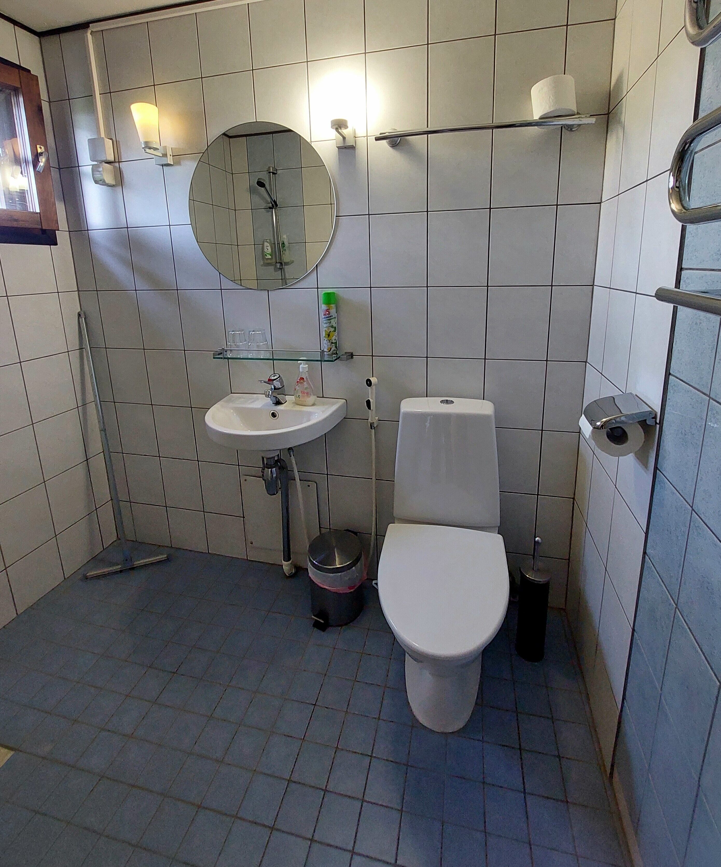A small, tiled bathroom with a white sink, mirror, toilet, and a trash bin.