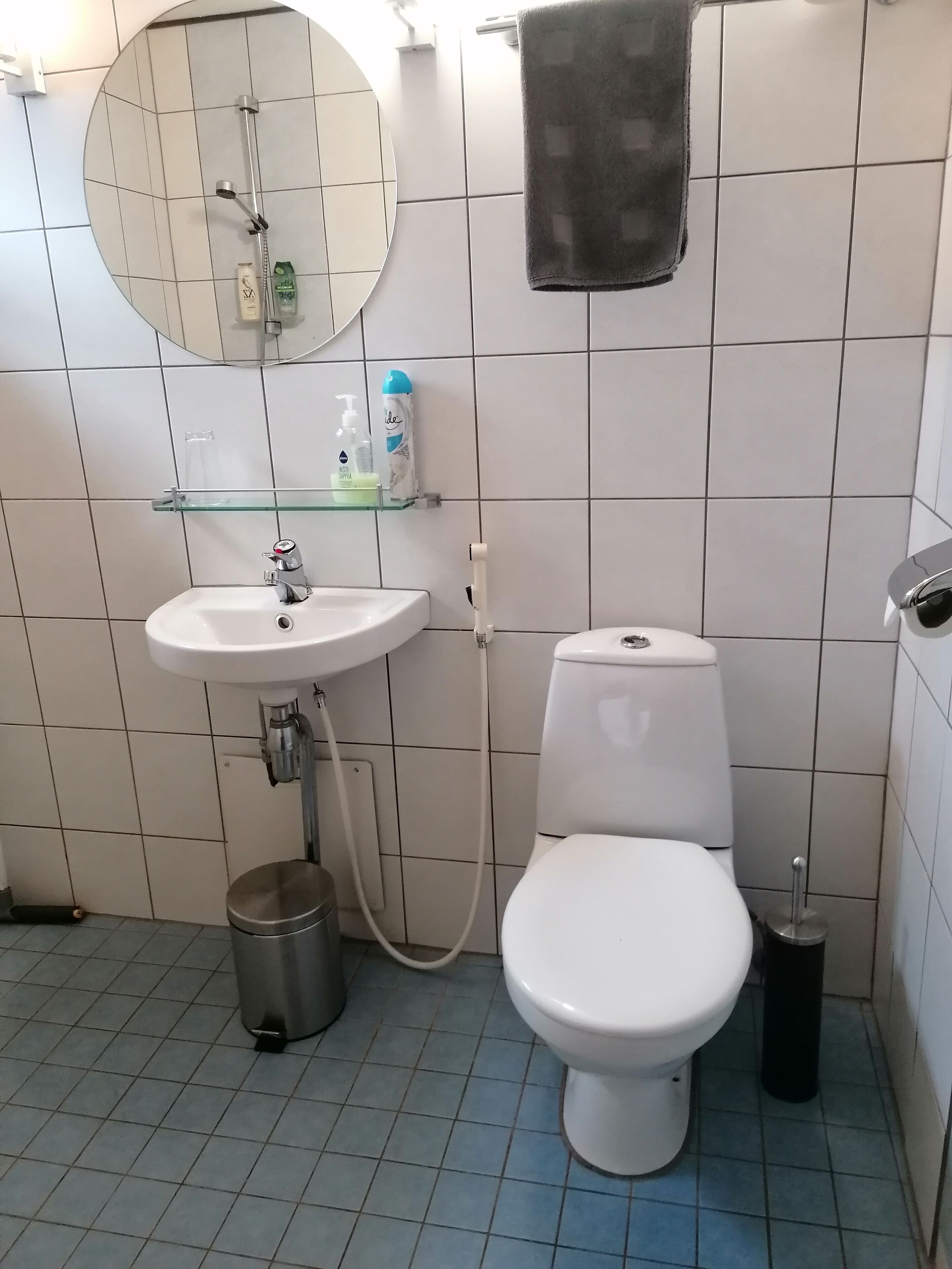 Un baño con lavamanos blanco, inodoro y un espejo redondo. Hay una toalla colgada en la pared, y se ve un bote de basura.