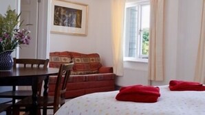 A room with a bed, a table, and a chair. There is a window with curtains, a framed picture on the wall, and a potted plant. - Royal Oak (Kingsbridge)
