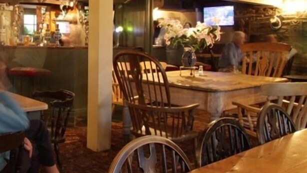 A restaurant interior with wooden tables and chairs, a bar area, and a television screen. - Royal Oak (Kingsbridge)