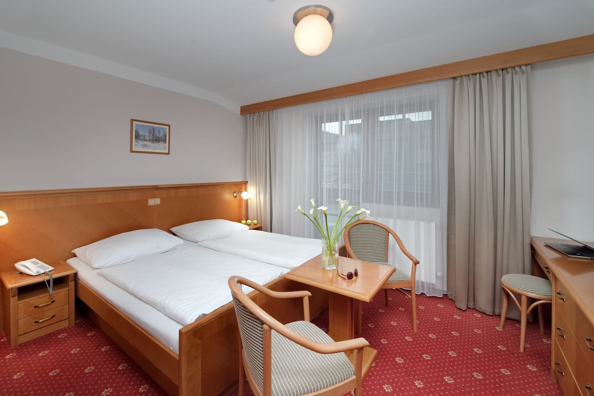 A hotel room with a large bed, a wooden desk, a chair, a small table, a nightstand with a phone, and a window with curtains.