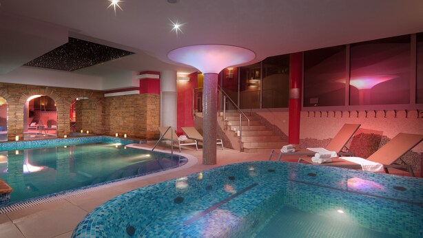A large indoor swimming pool with tiled walls and a central column, surrounded by lounge chairs and a staircase. - Wellness Hotel Windsor (Spindleruv Mlyn)