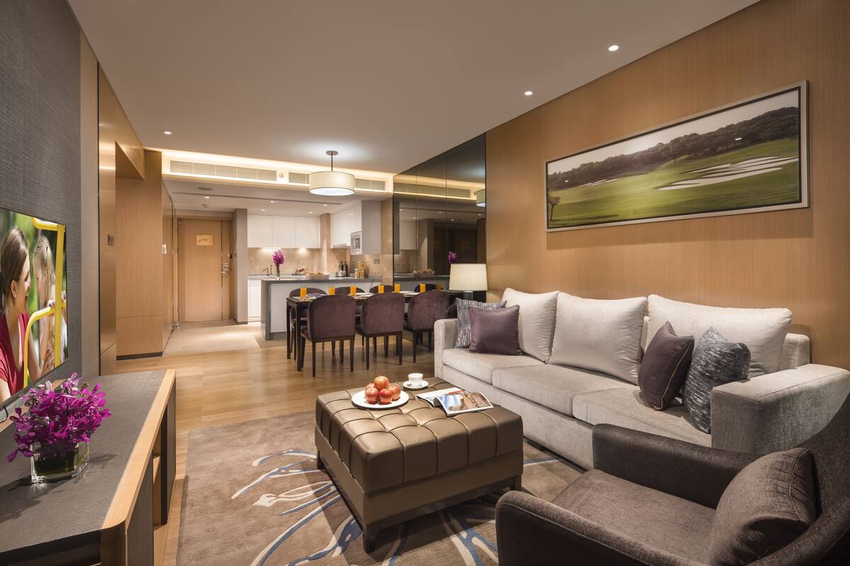 A modern living room with a sofa, ottoman, and chairs, a dining area with a table and chairs, and a kitchen in the background.
