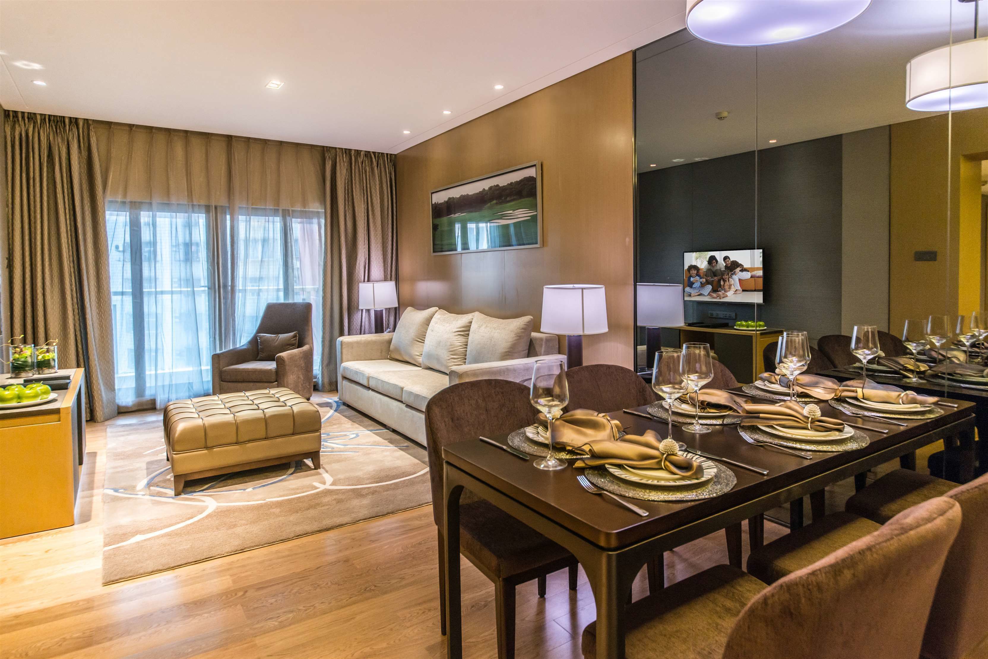 A modern hotel room with a dining area, a sofa, a television, and a desk with a lamp.