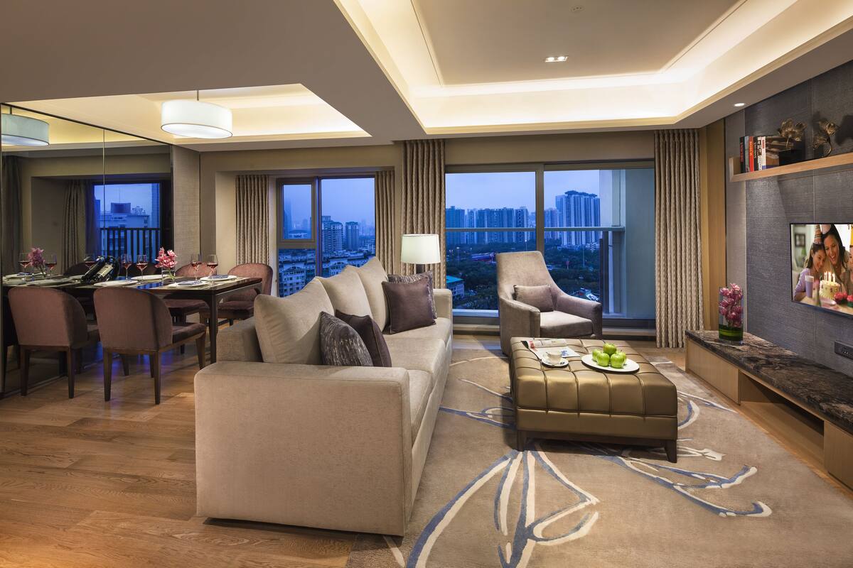 A modern hotel room with a sofa, armchair, dining area, and a large window with a city view.