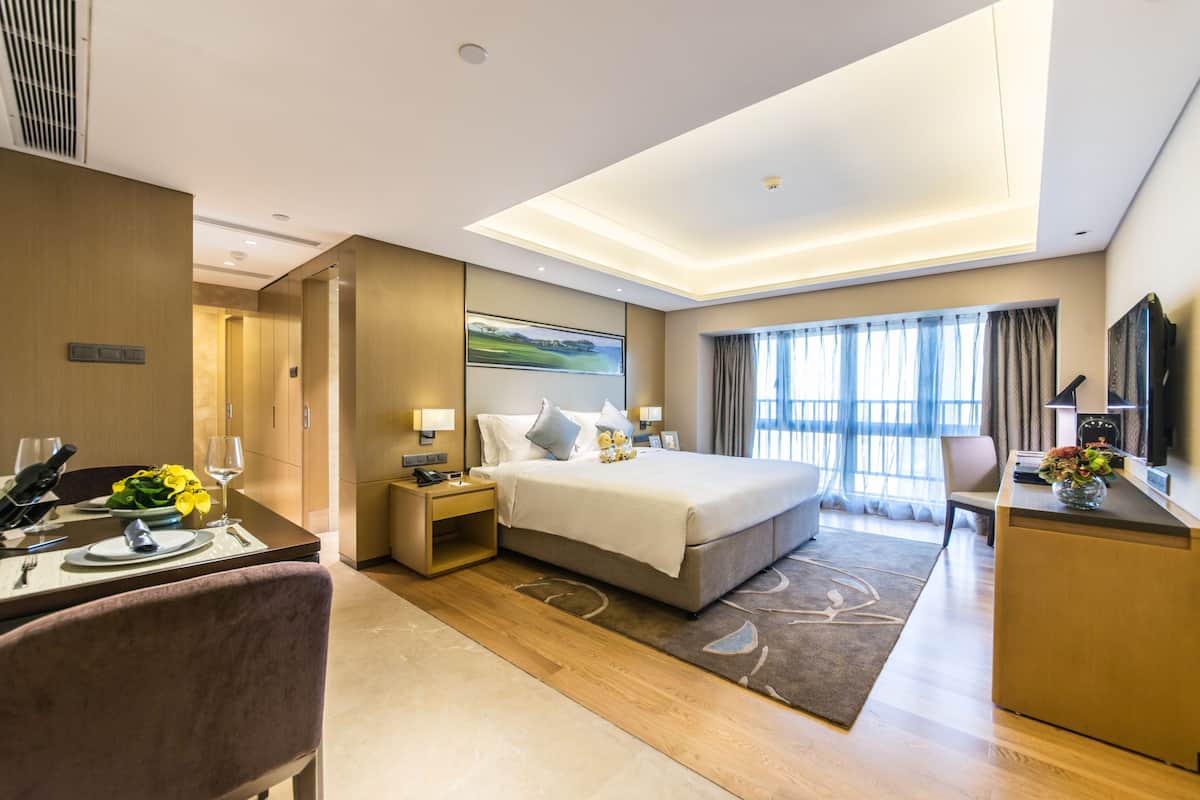 A modern hotel room with a large bed, a desk with a chair, a desk lamp, a television, and a painting on the wall.