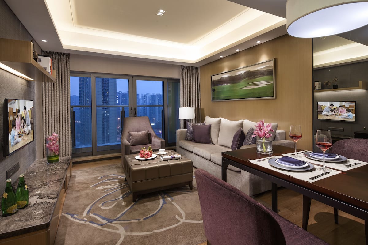 A modern hotel room with a large window, a sofa, a dining table set for two, and a television on the wall.