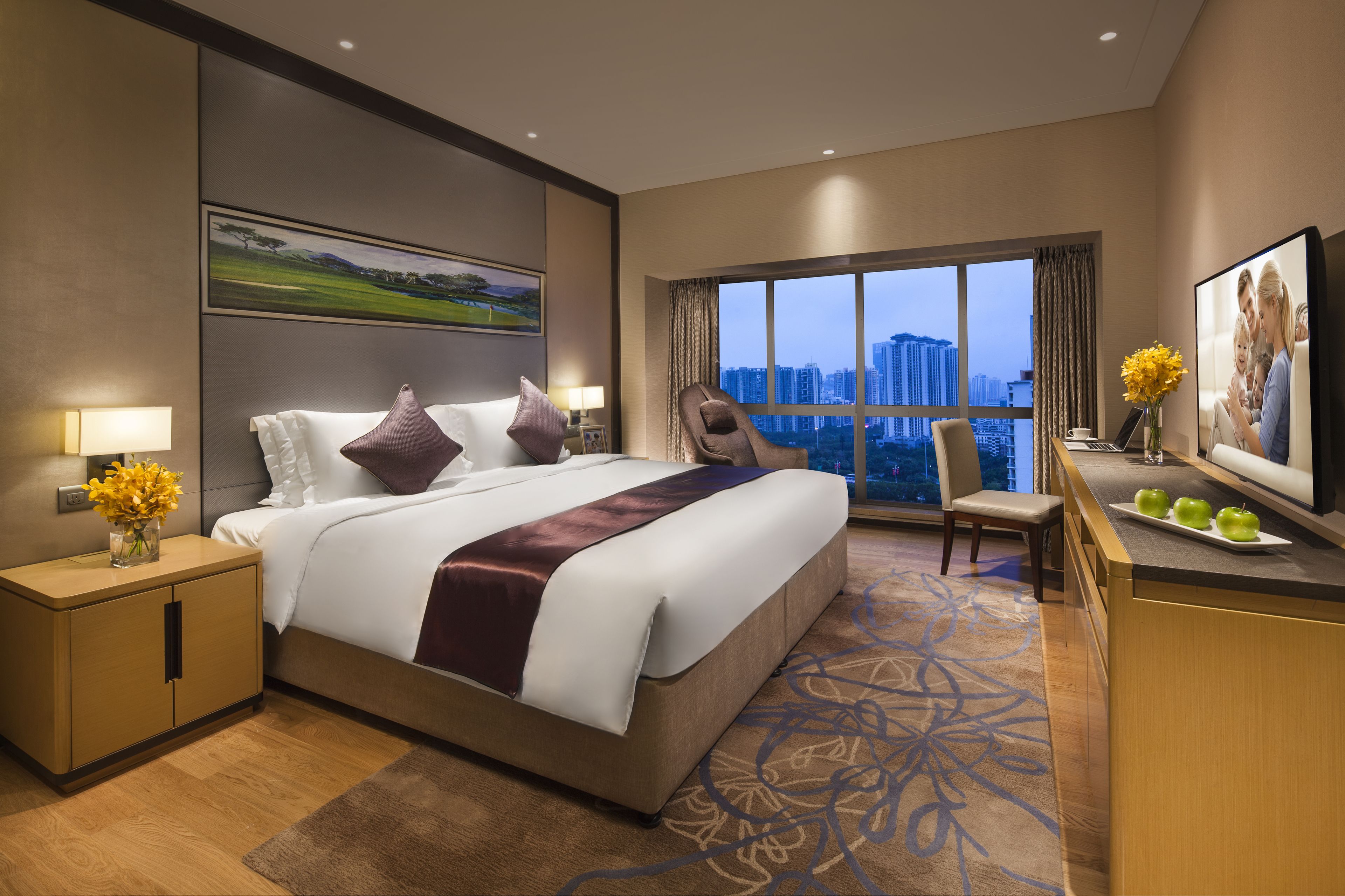 A hotel room with a large bed, a desk with a laptop, a chair, and a view of the cityscape through large windows.