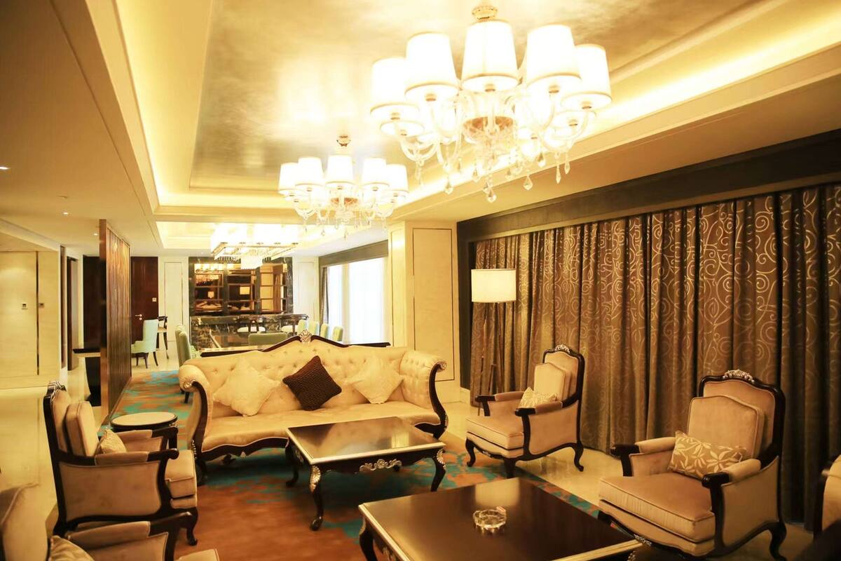 Un salon spacieux, doté d’un canapé, de fauteuils et d’une table basse. Un lustre est suspendu au plafond et des rideaux ornent les fenêtres.