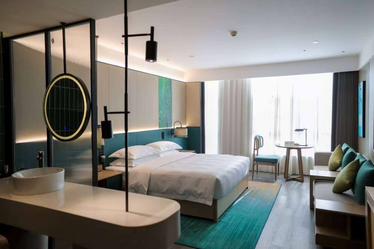 Une chambre d’hôtel moderne avec un grand lit, un miroir rond, une table de chevet, un petit bureau et une vue sur l’extérieur grâce à de grandes fenêtres.
