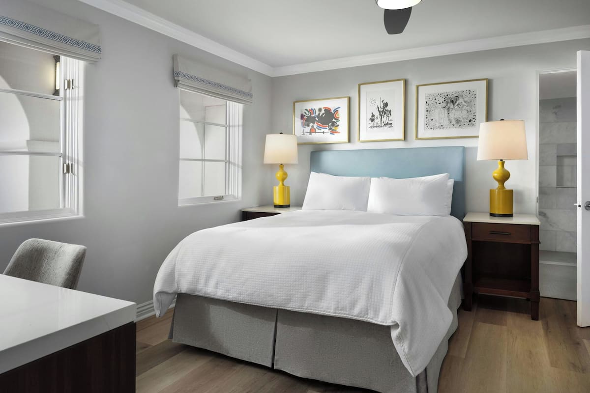 A hotel room with a large bed, two bedside tables with lamps, a desk, and a chair. There are three framed pictures on the wall above the bed.