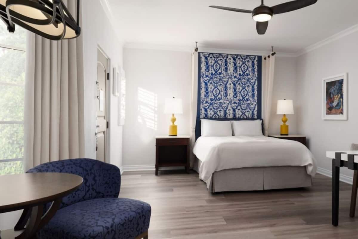 A bedroom with a bed, a chair, a table, a ceiling fan, and a nightstand with a lamp.