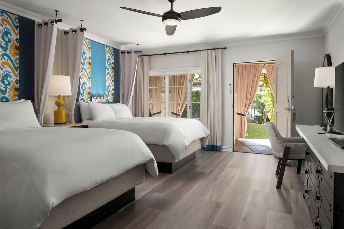 A hotel room with two beds, a ceiling fan, a desk with a lamp, and a view of the outdoors.