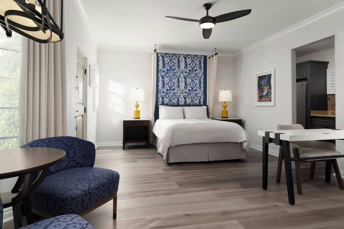 A hotel room with a bed, a desk, a chair, a table, a ceiling fan, and a window with curtains.