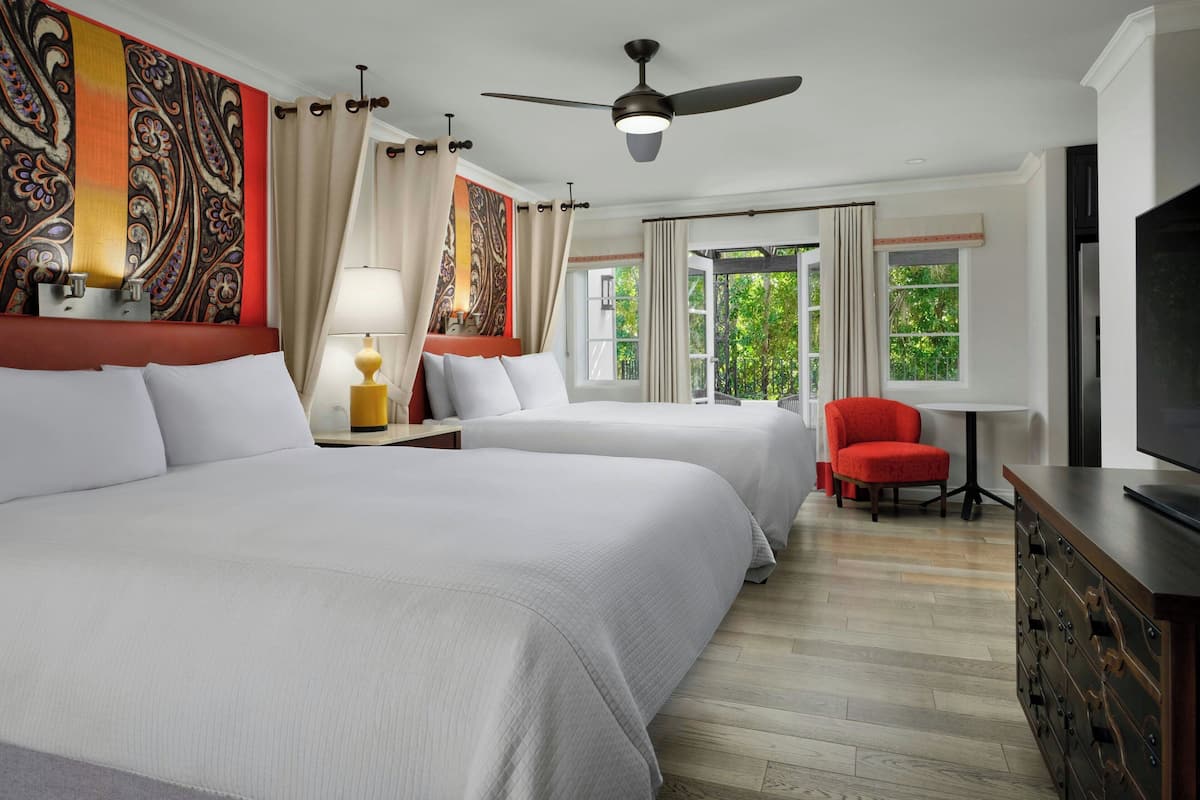A hotel room with two beds, a ceiling fan, a TV, and a view of greenery outside.