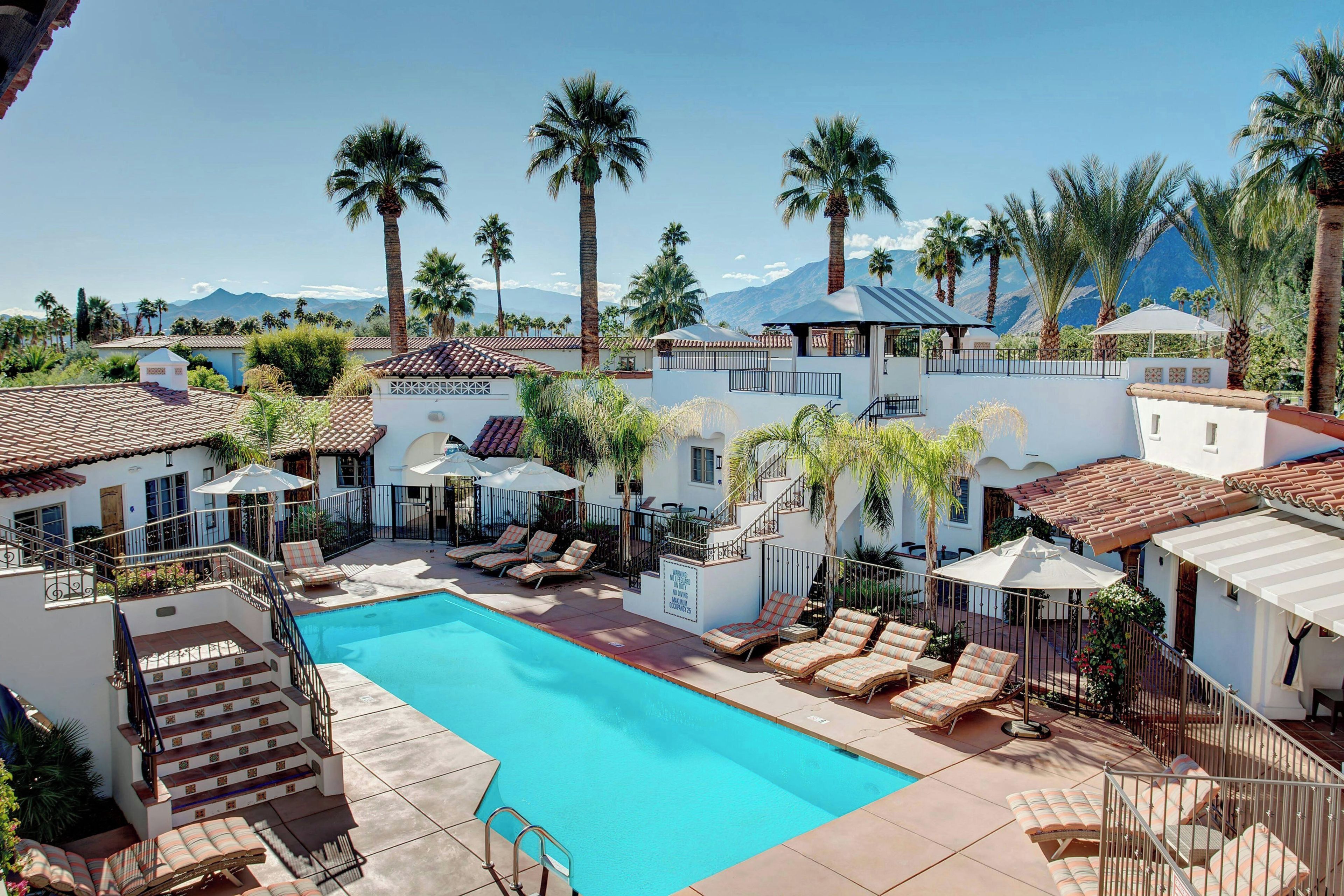 A pool area with lounge chairs and umbrellas, surrounded by palm trees and a mountain view.