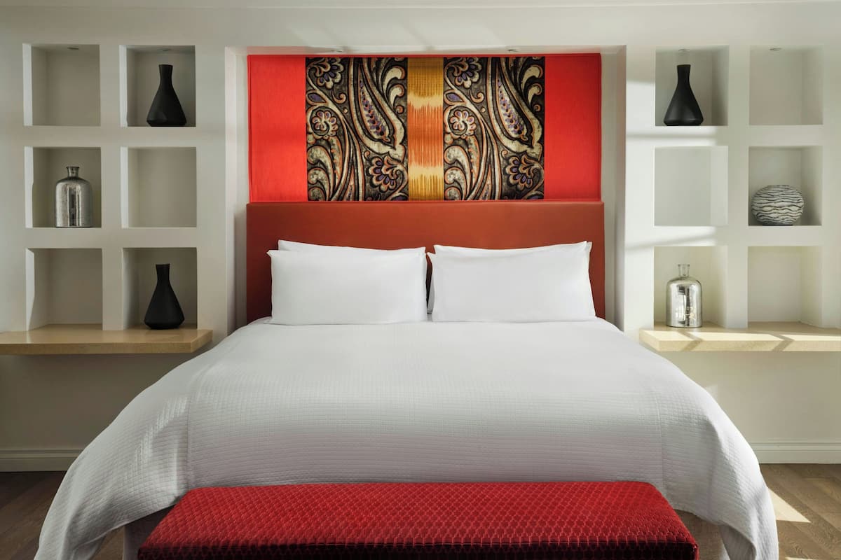 A modern hotel room with a large bed, white bedding, a red accent wall, and shelves with decorative items.